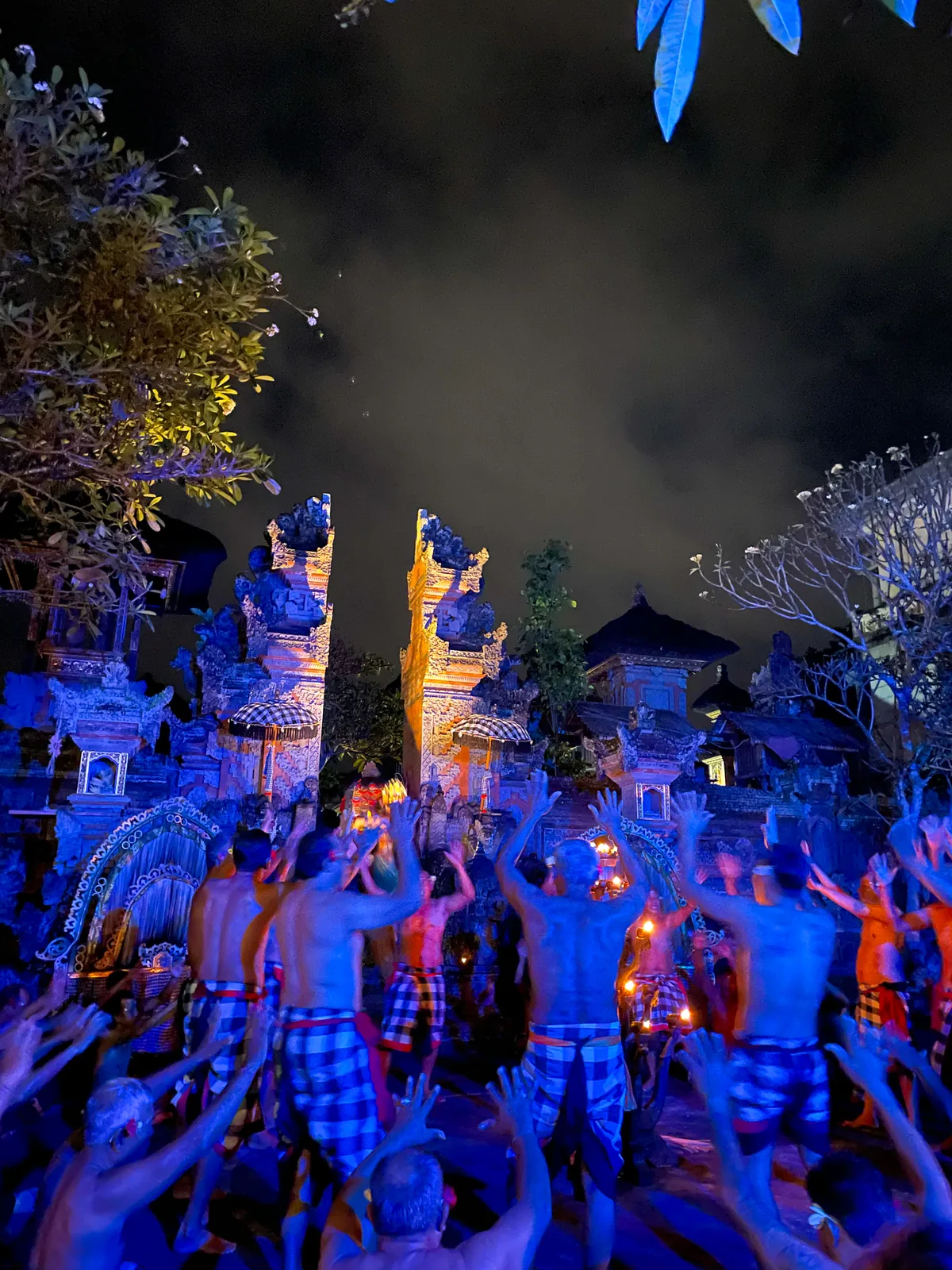 Kecak dance at night, chanting men in checked sarongs before a temple gate lit blue