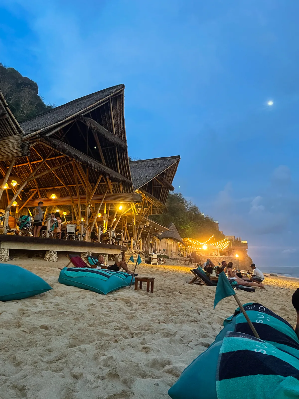 The bamboo bar at Sundays Beach Club lit up at dusk, teal beanbags on the sand