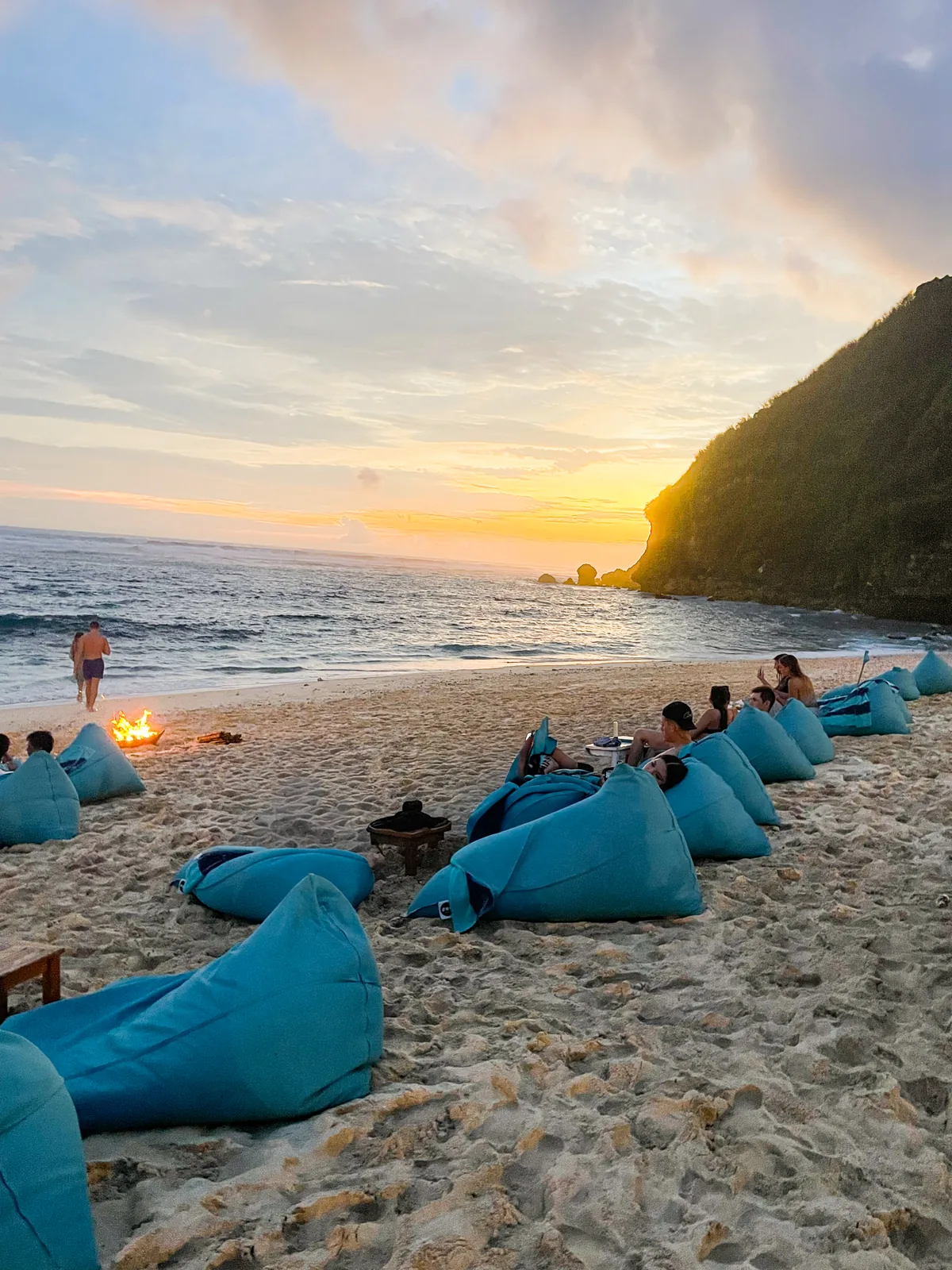 Rows of teal beanbags and a small bonfire on the sand at Sundays Beach Club at sunset, below a green headland