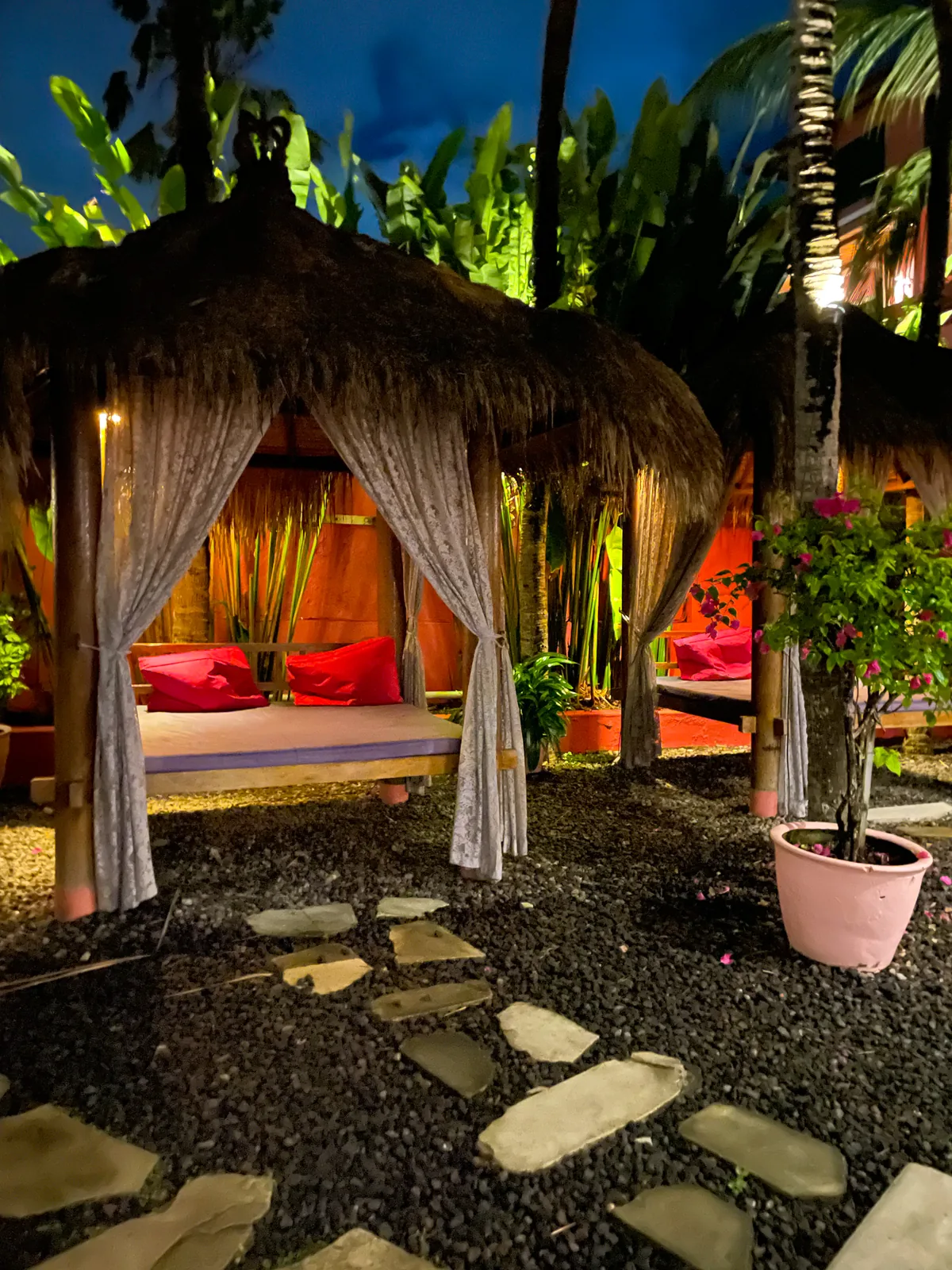 Thatched garden cabana with curtains and red cushions, lit up at night