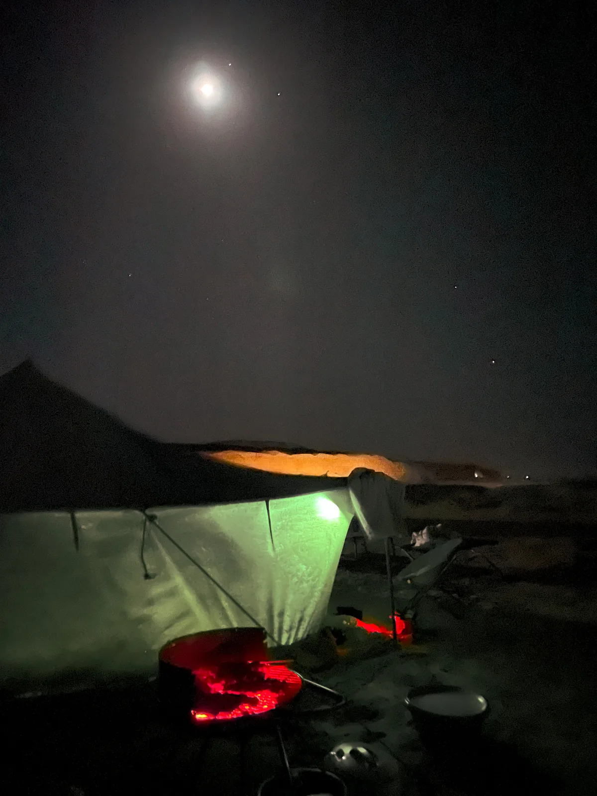 Night at camp: a bright moon above a tent lit green from inside, glowing charcoal on the barbecue and camp chairs in the foreground