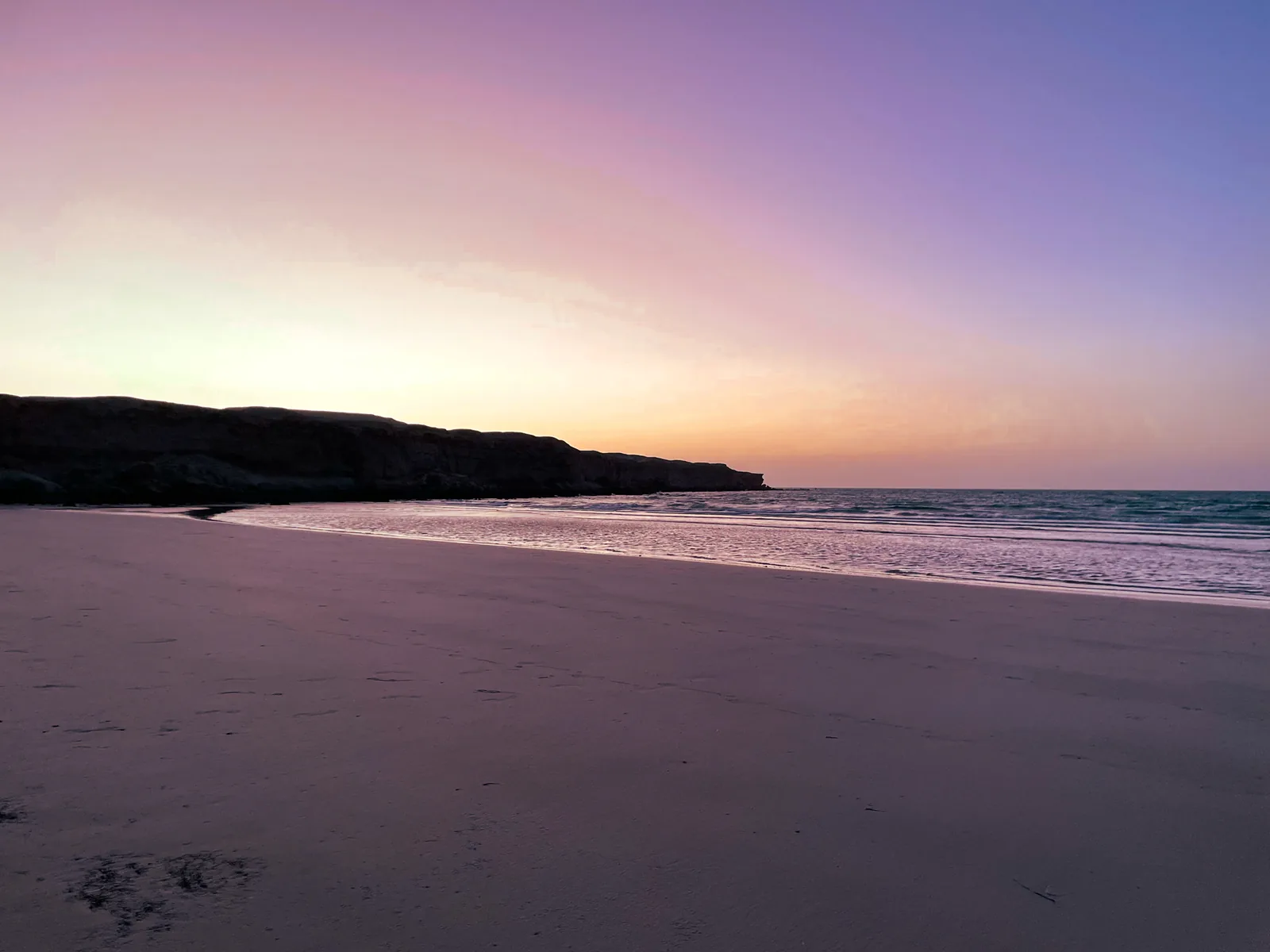 Pink and lilac sky after sunset over a flat empty beach and calm sea, with a dark cliff headland on the left