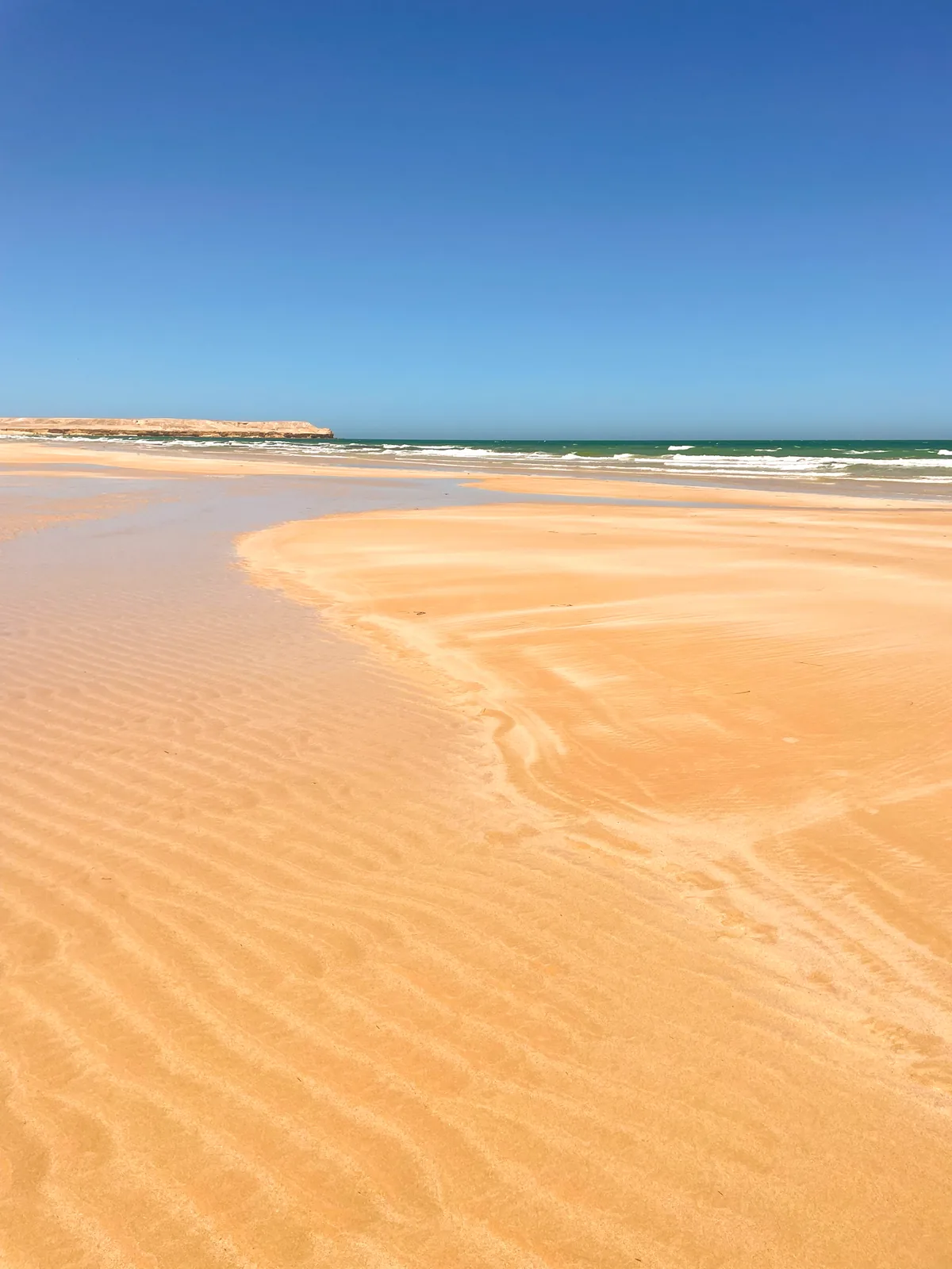 Ripples in orange sand at low tide beside a shallow channel, with white surf and a flat-topped headland under a deep blue sky