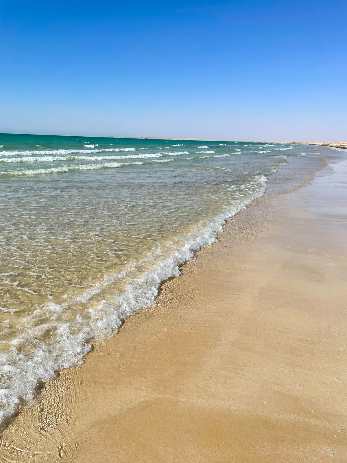 Small turquoise waves washing onto golden sand along a long, straight, empty shoreline under a clear blue sky