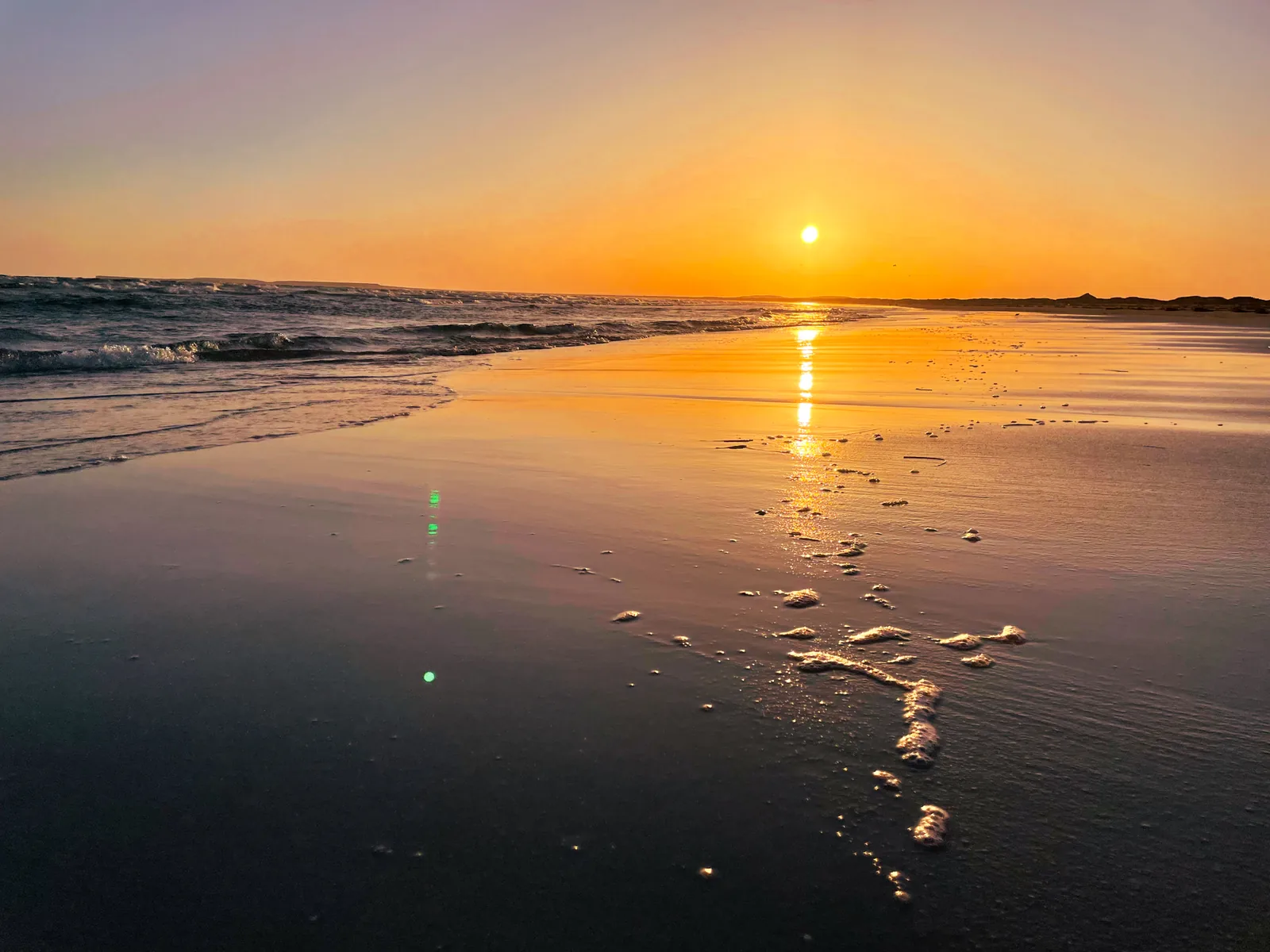 An orange sunset sky reflected in a streak of light across wet sand, with sea foam in the foreground