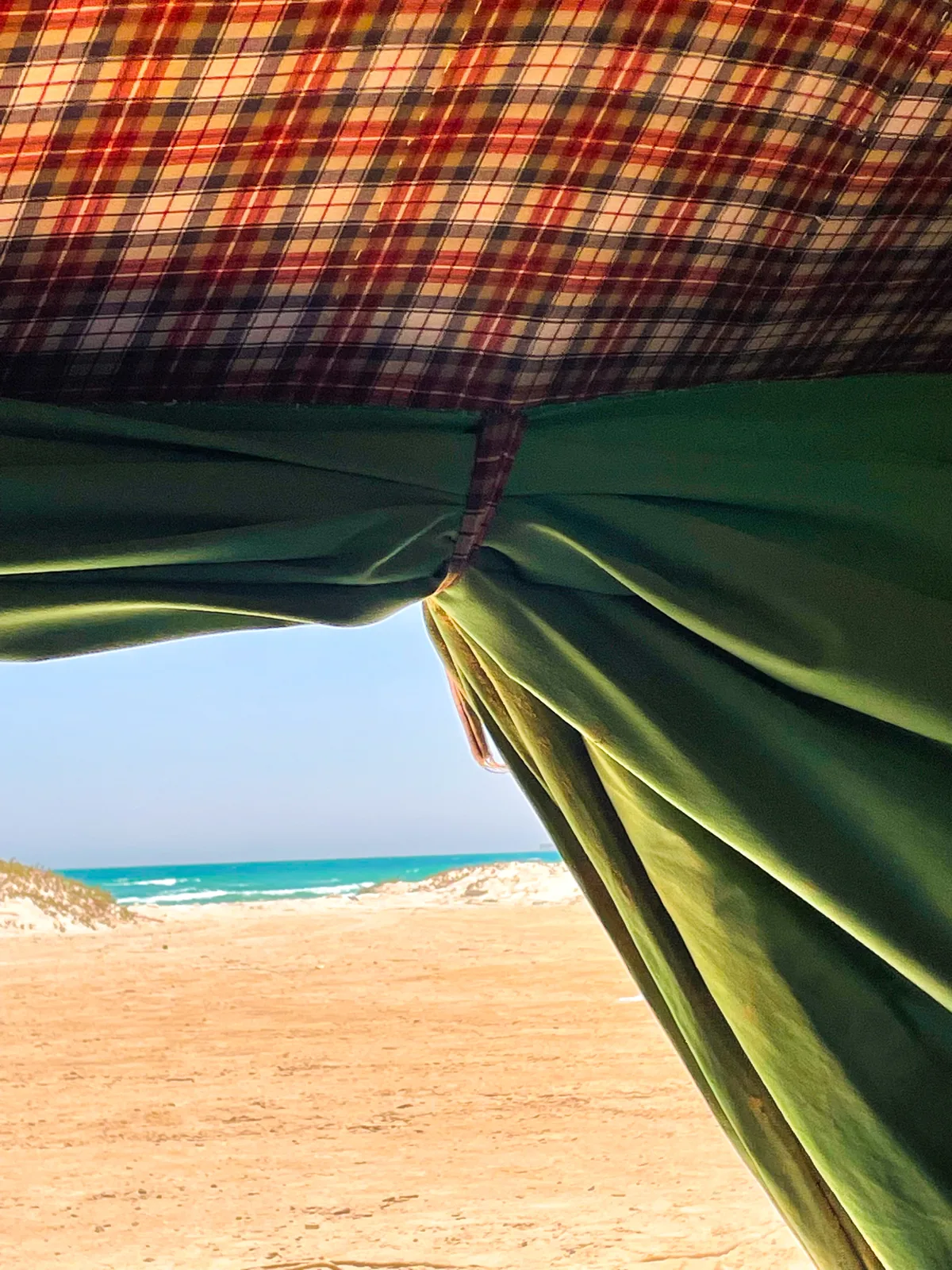 The view from inside the tent: plaid lining overhead and a green wall pulled back to frame sand, dunes and turquoise sea