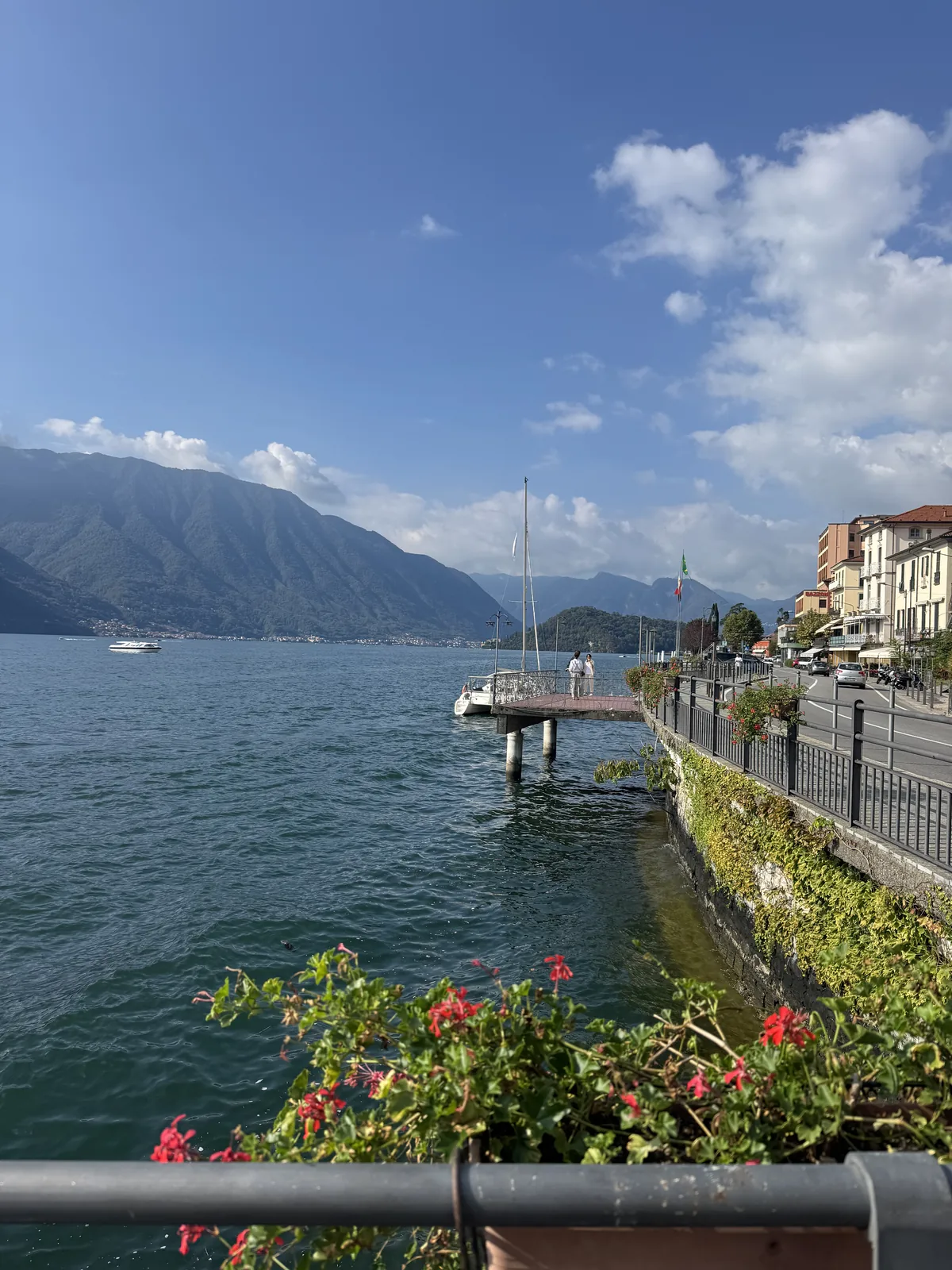Lake Como dock scene with a small sailboat moored, red flowers in the foreground and mountains across the water