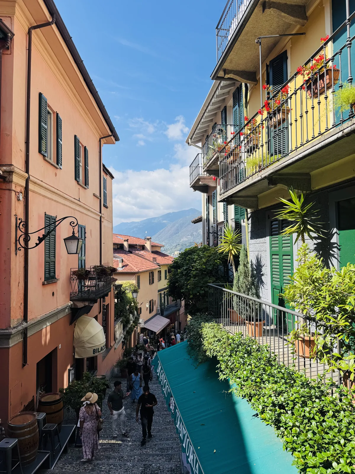 Bellagio's steep, colourful stepped alley with balconies of red geraniums and tourists walking down toward the lake