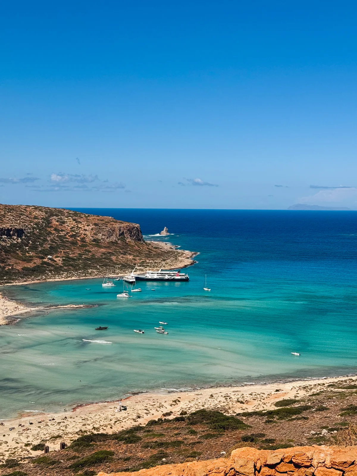 Balos bay from the path, a cruise ferry anchored among small boats and yachts beside a rock pinnacle