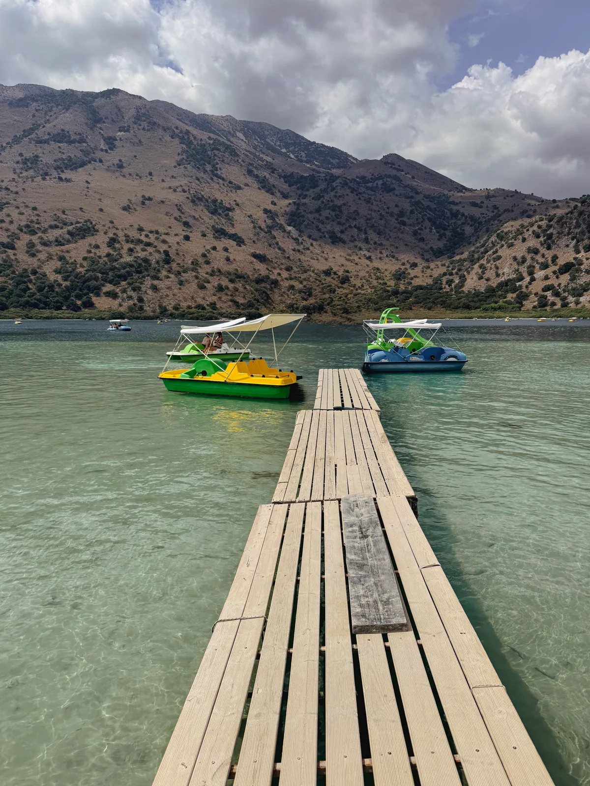 Wooden jetty leading into clear green water at Lake Kournas, pedalos moored and bare mountains behind