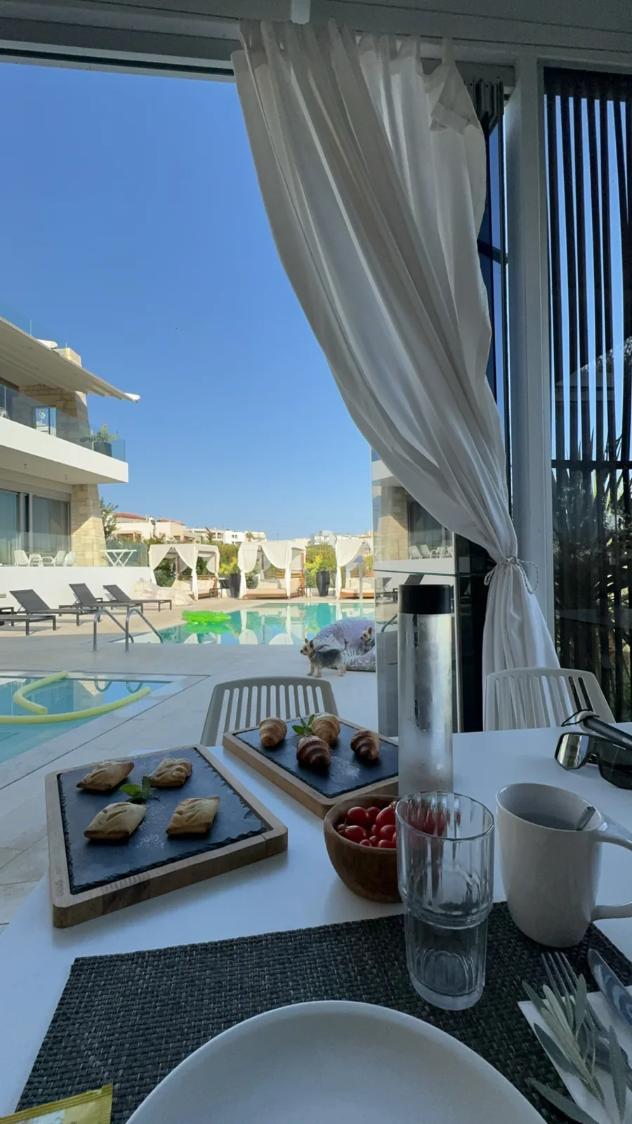 Breakfast table by the pool with slate boards of pastries and tomatoes, the pool and cabanas behind