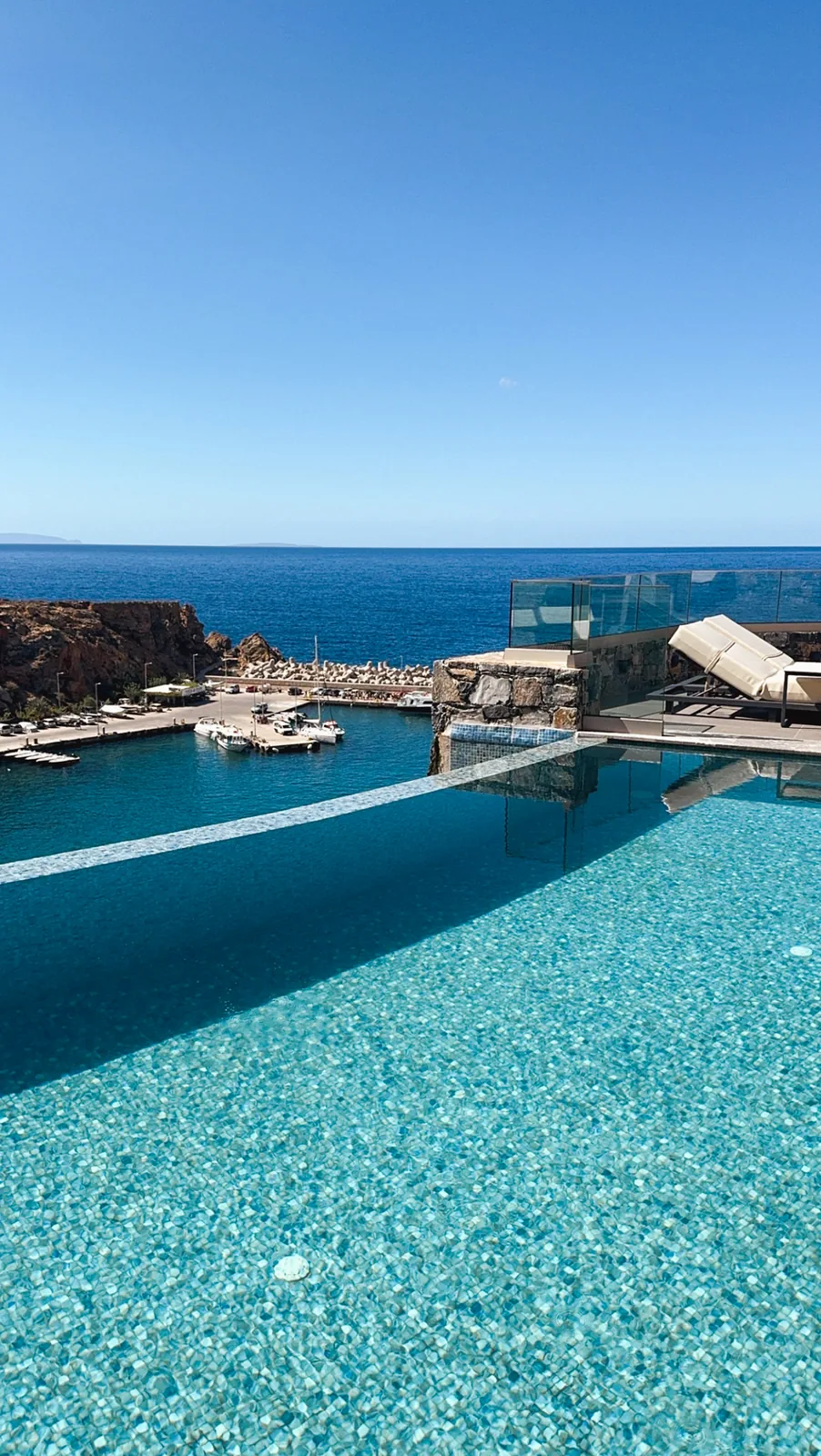 The edge of the infinity pool above the small port of Chora Sfakion, with a glass balustrade and a sunbed