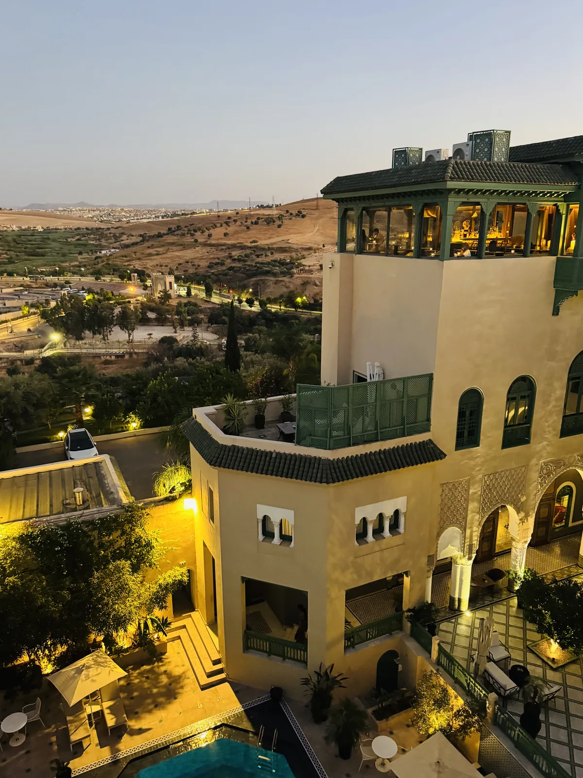 Palais Faraj at dusk, its lit rooftop bar above the pool with the hills of Fes beyond