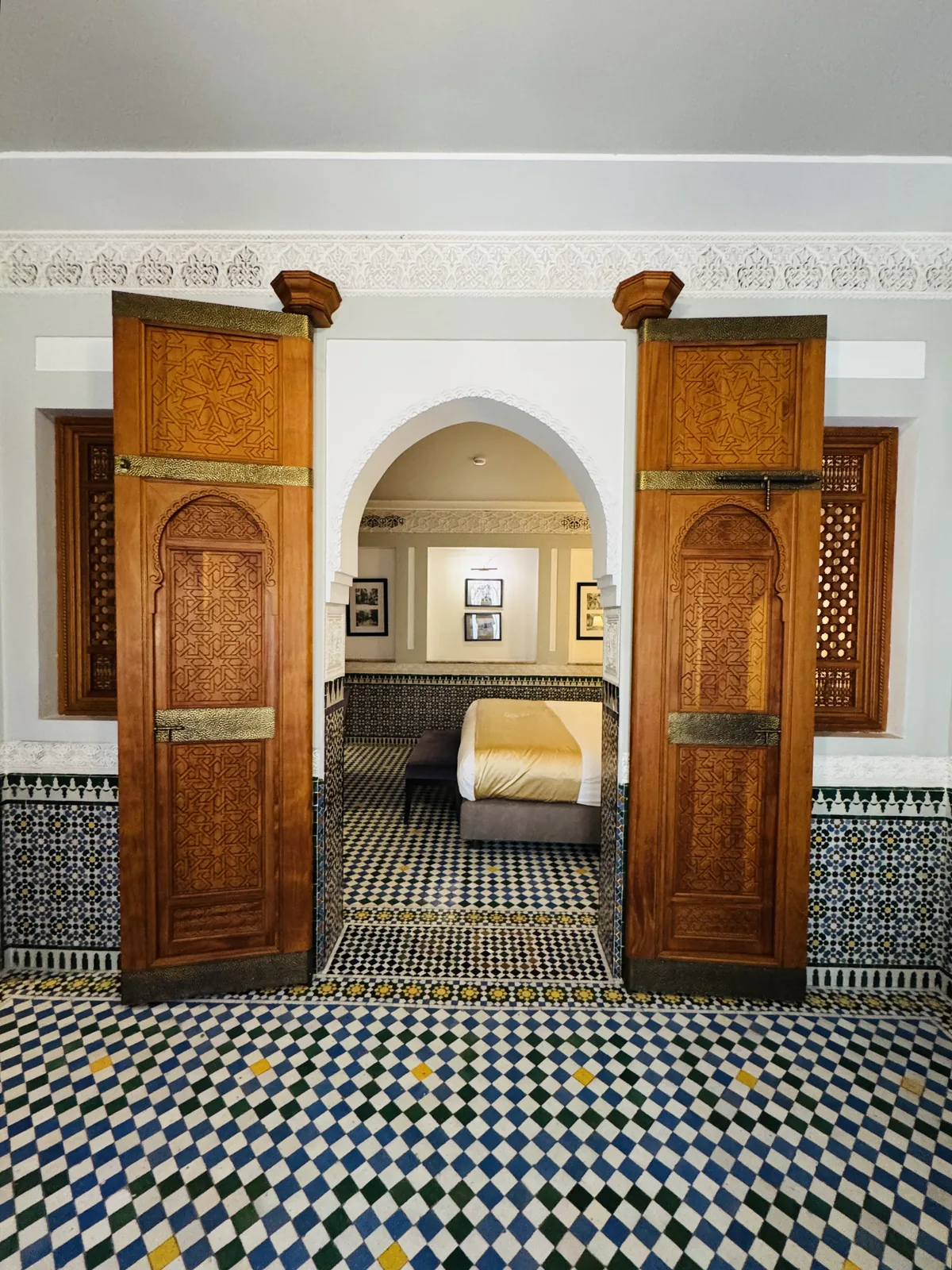 A riad room in Fes with carved cedar doors and zellige tiling, a bed glimpsed through the arch