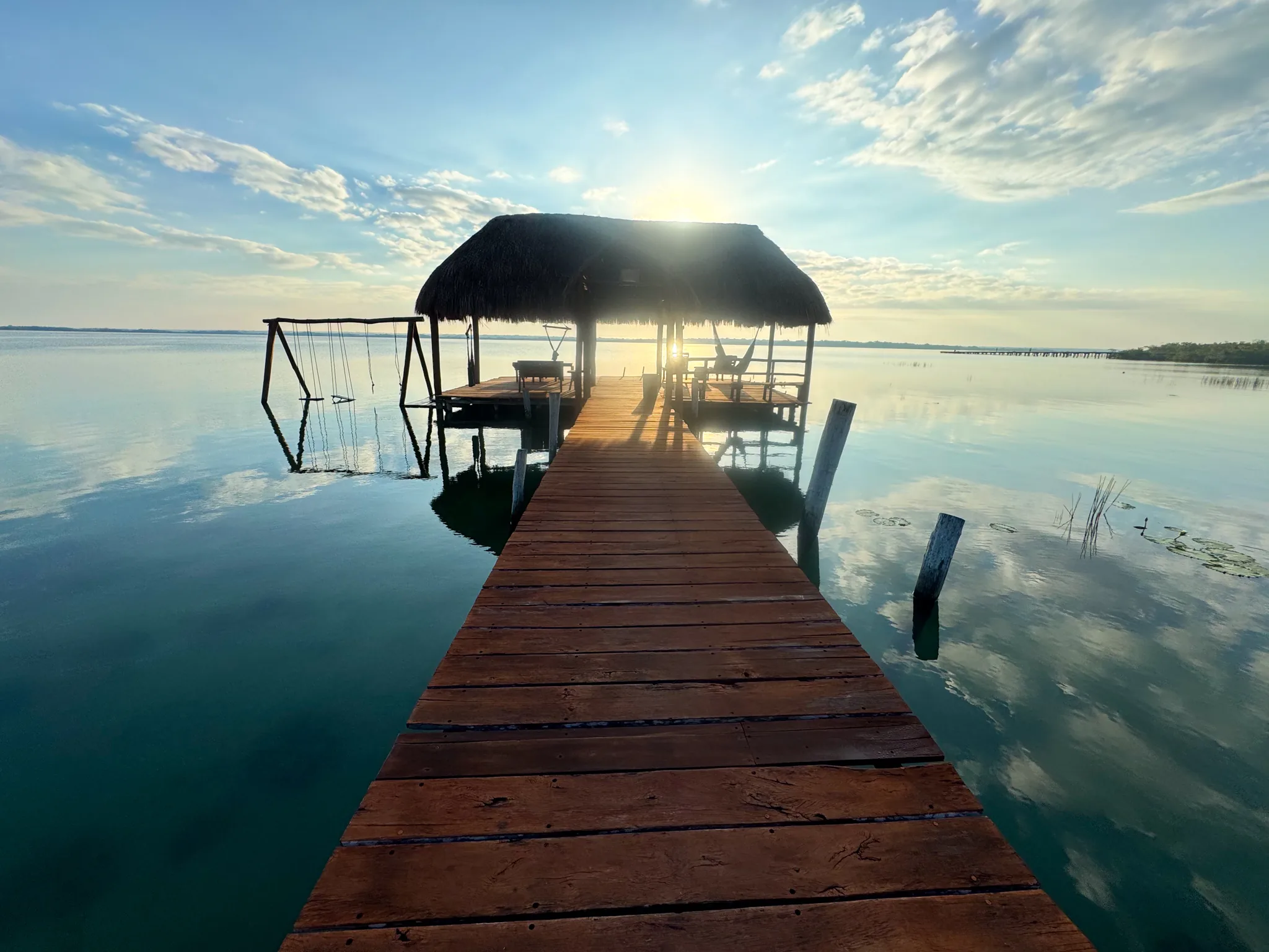 A wooden dock leading to a thatched palapa on the Bacalar lagoon at sunrise