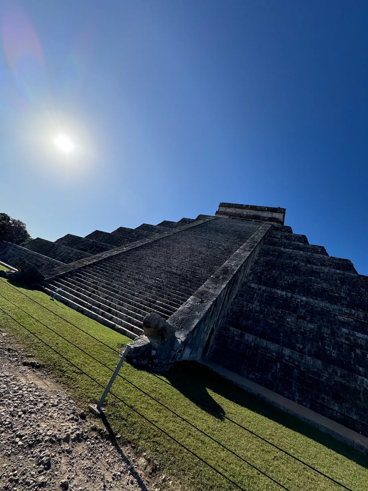 Low angle along the steps of El Castillo, the carved serpent head at its base, with a sun flare
