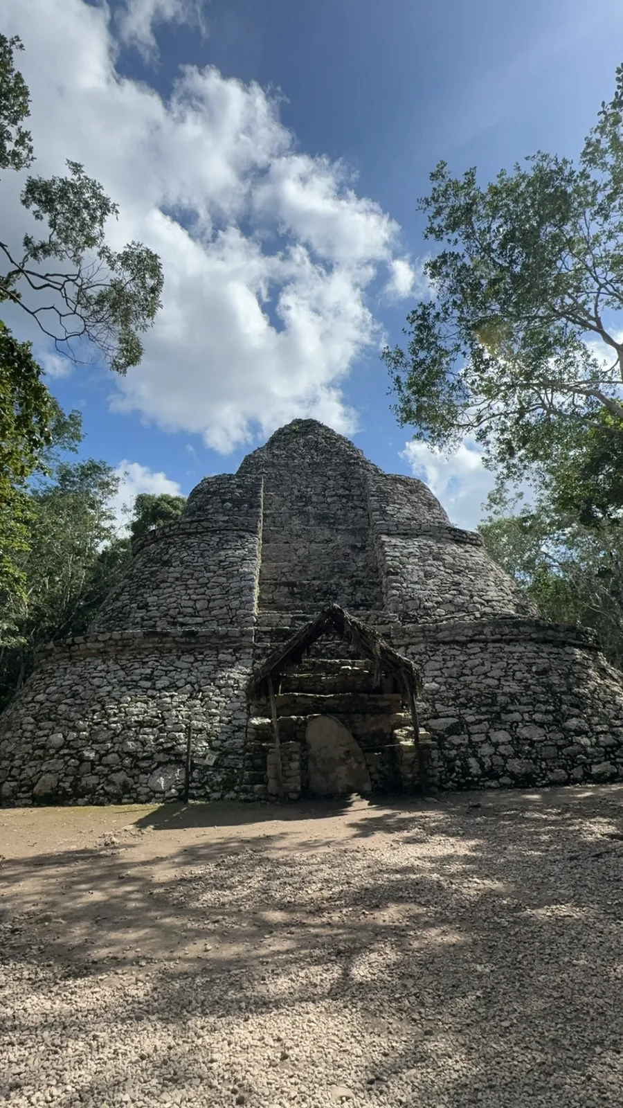 A rounded stepped stone structure at the Cobá ruins with a thatched shelter under clouds