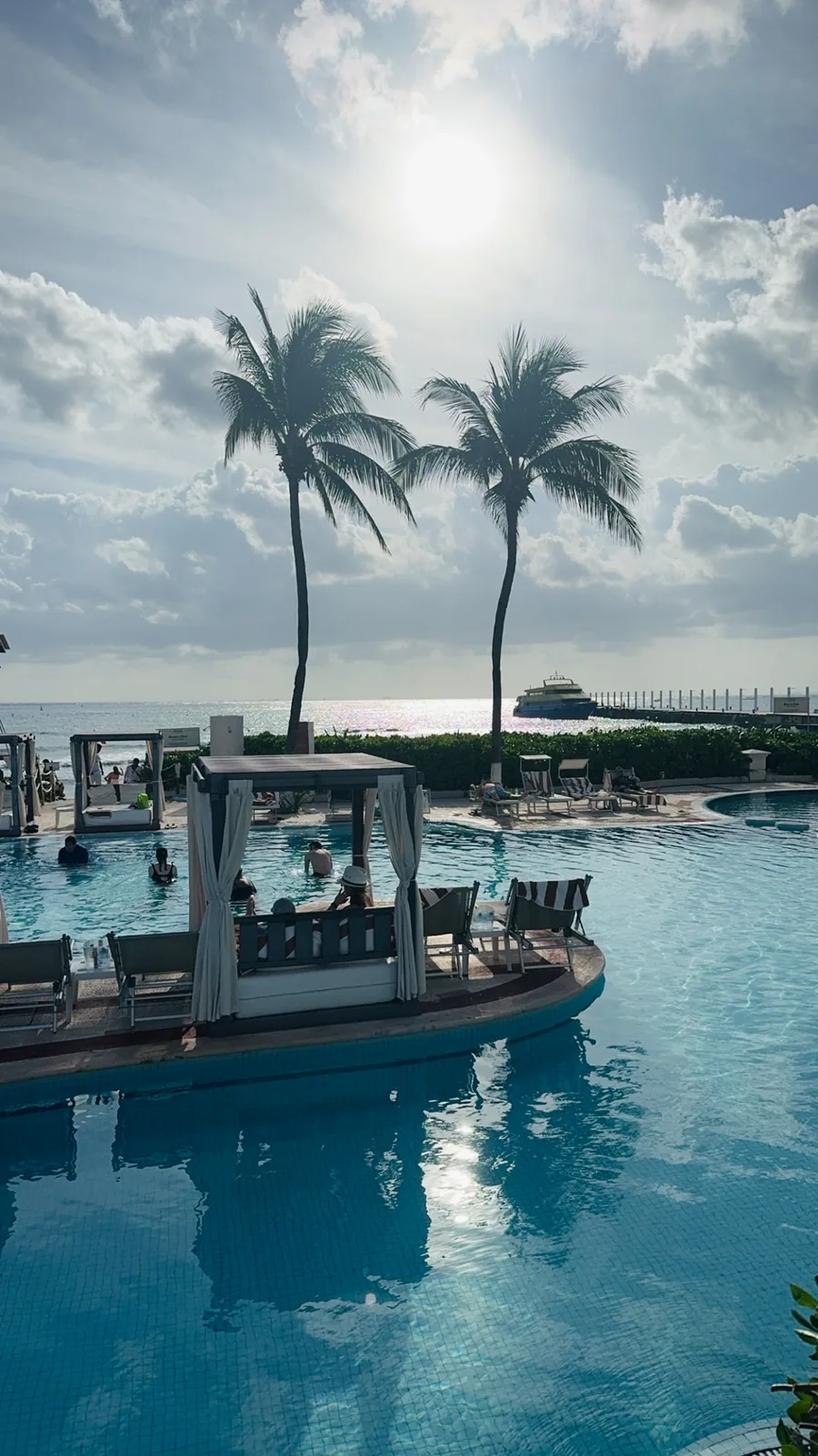 Hilton Playa del Carmen pool with a curtained cabana and two palms, a ferry at the pier behind