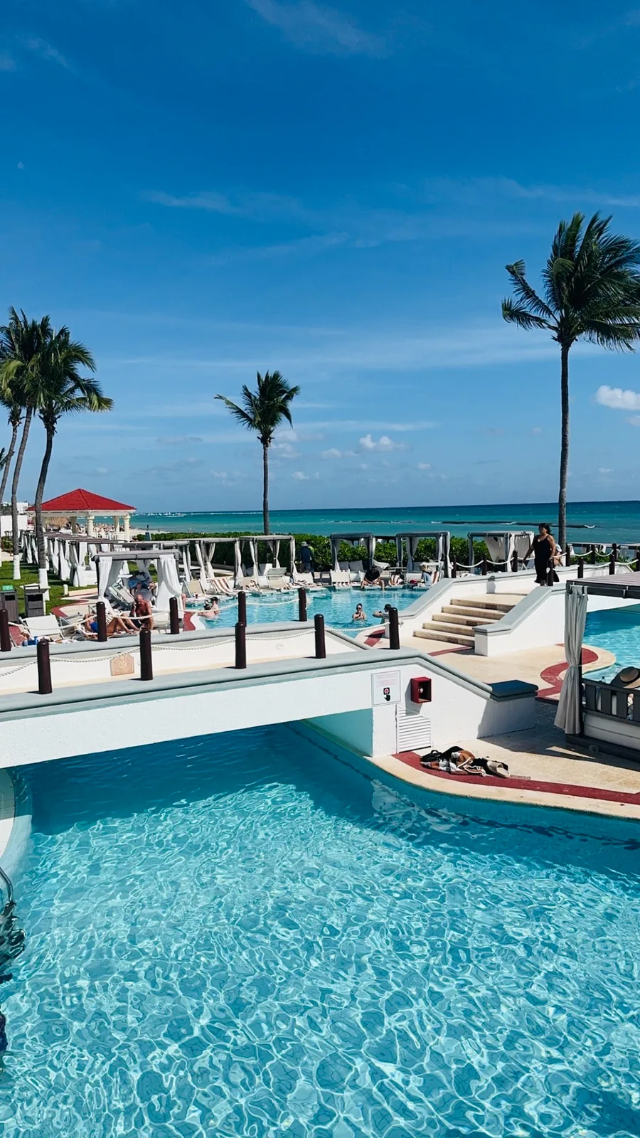 Blue pools linked by a small bridge at the Hilton Playa del Carmen, loungers, palms and the sea beyond