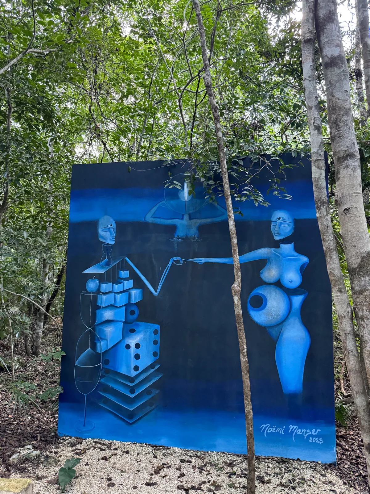 A large blue surrealist painting standing among the trees on the Holistika Art Walk